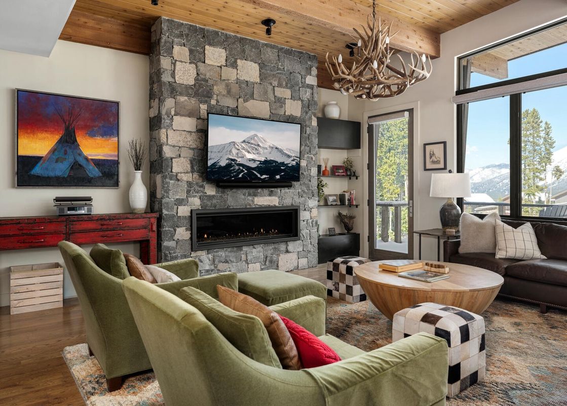 A luxurious living room featuring a stone fireplace, antler chandelier, green armchairs, and a large window with mountain views.