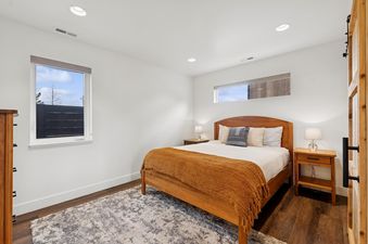 A modern bedroom with a wooden bed frame, an orange-brown throw blanket, two nightstands with lamps, and two windows with an outdoor view.