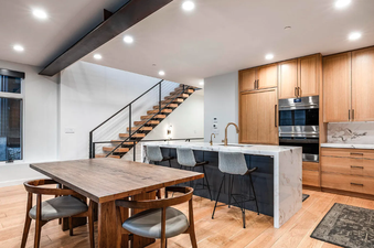 A modern kitchen and dining area with a wooden dining table and chairs in the foreground, a kitchen island with bar stools, and a wooden staircase in the background.