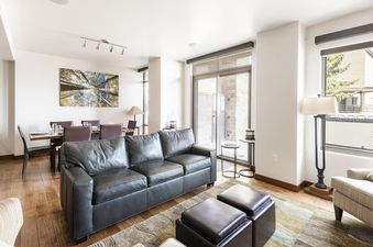 A spacious and inviting open-concept living and dining area at Lowell 101, featuring a dark leather sofa, two matching ottomans, and a dining table with chairs, all illuminated by ample natural light.