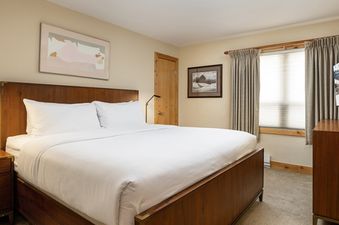 The Montaneros 202 bedroom features a king-sized bed with a dark wood frame, crisp white linens, and decorative artwork, alongside a window with blinds and drapes.