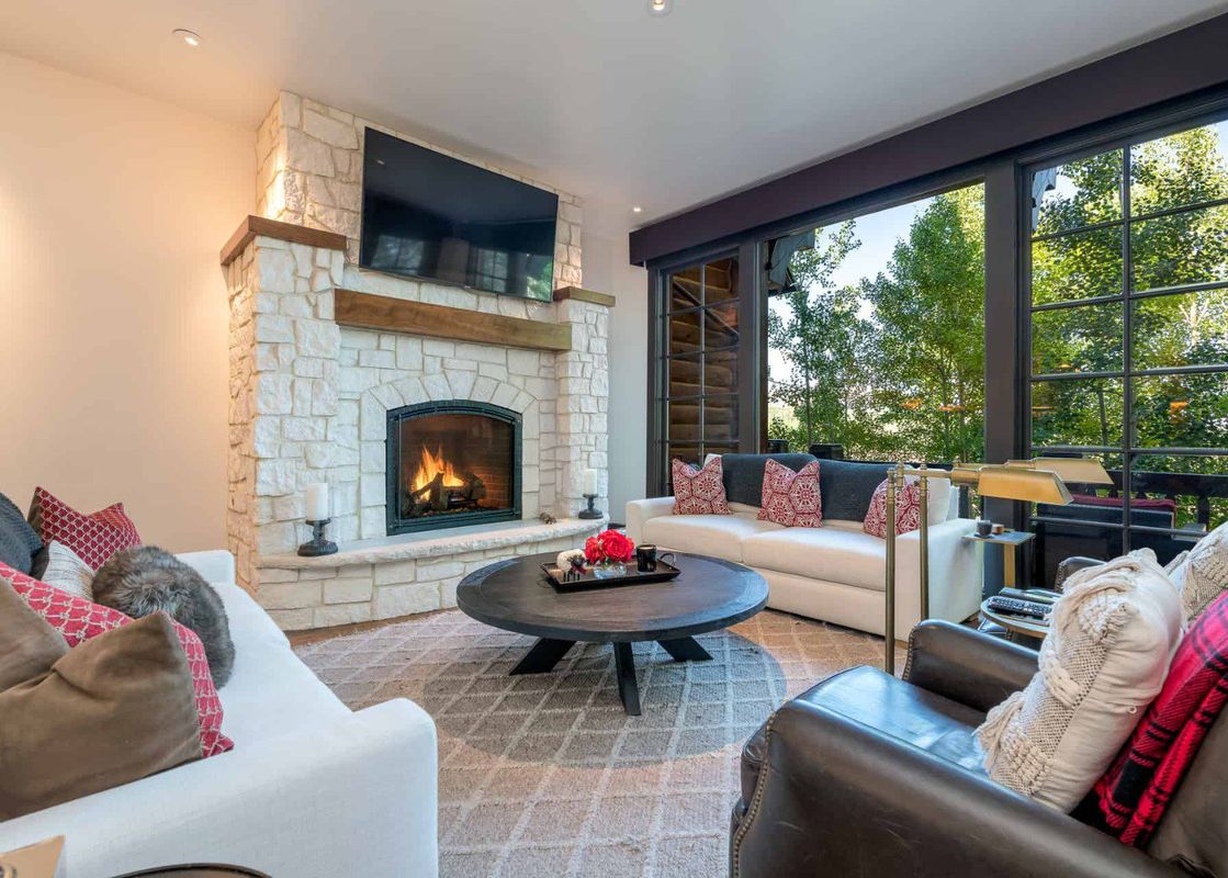 A living room with a fireplace, two white couches, and a large window overlooking a lush green forest.
