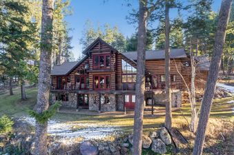 A large, rustic log cabin with deep red window frames and a wrap-around balcony, nestled among tall pine trees with a view of a snowy, grassy landscape.