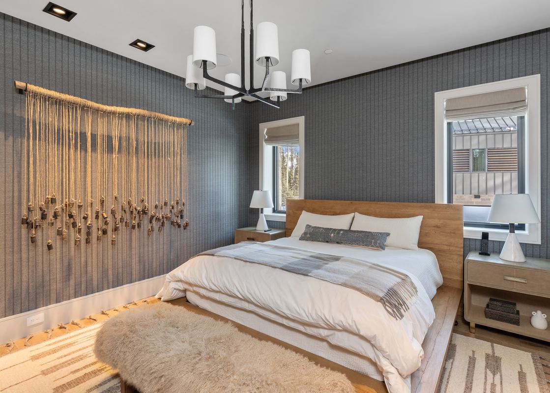 A cozy bedroom features a king-size bed with light wood headboard, white bedding, and a plaid throw blanket, flanked by two nightstands with lamps, a decorative rope and bell wall hanging on patterned grey wallpaper, and a window with blinds offering a gl