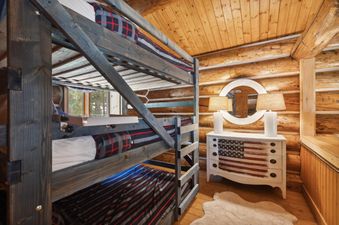 The bunk room at Apres Chalet features wooden bunk beds with plaid and striped bedding, rustic log walls, and a dresser with an American flag design under a round mirror.