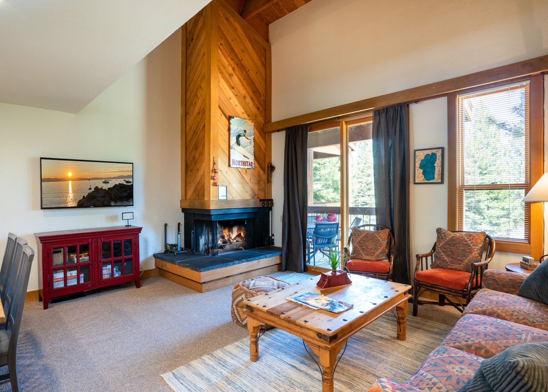 The Northstar Gold Bend Getaway's spacious living room features a lit fireplace, a large flat-screen TV displaying a sunset, comfortable seating, and access to a balcony through a sliding glass door.