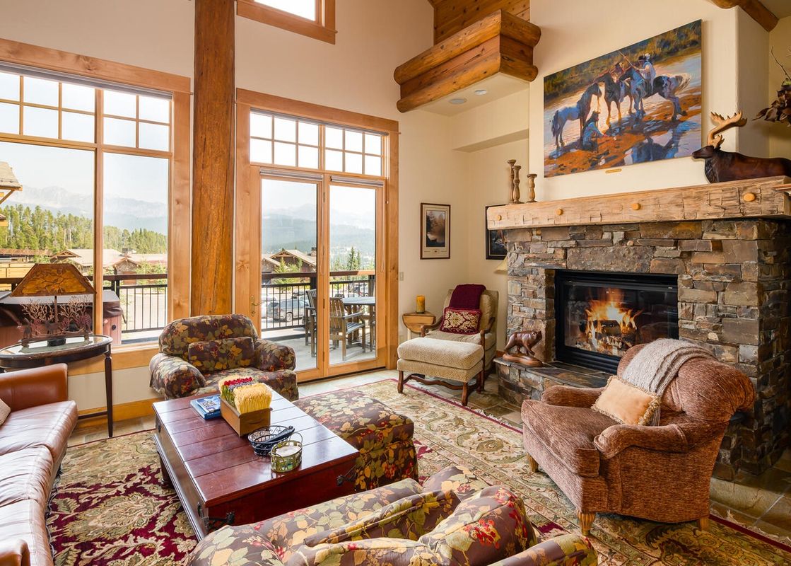 The living room of Moonlight Basin Penthouse 4 has a large stone fireplace with a roaring fire, large windows with views of the mountains, and comfortable seating.