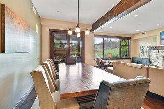 A modern dining room with a large wooden table and chairs, with views of trees from the windows.