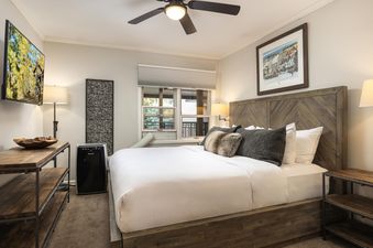 A cozy bedroom with a king-sized bed, wooden headboard, white bedding, decorative pillows, bedside tables, and a flat-screen TV.