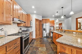 A fully equipped kitchen with wooden cabinetry, granite countertops, and stainless steel appliances, featuring a gas range and a large refrigerator.