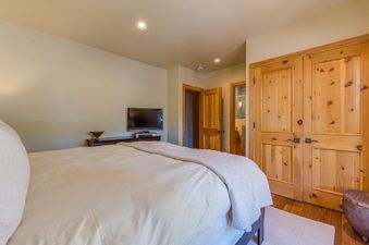 A cozy bedroom at See Forever Residence 110 features a bed with white and cream bedding, a wall-mounted TV, and wooden doors leading to other rooms, including a visible bathroom.