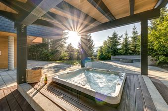 An inviting outdoor hot tub with bubbling water, set on a wooden deck under a covered patio, surrounded by lush green trees and bathed in bright sunlight.