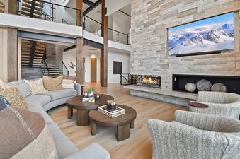 The great room at the Deer Crest Escape Retreat features a grand stone fireplace with a TV above, plush curved sofas, wooden coffee tables, and a sweeping staircase.