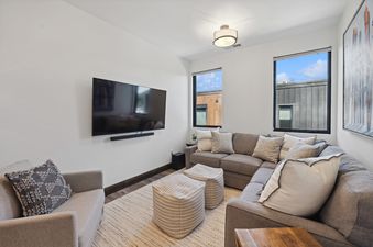 A modern media room with a large TV, a comfortable sectional sofa with throw pillows, two striped ottomans, and two windows offering an exterior view.