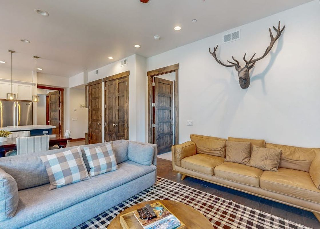 The living room of Modern Blackstone Paradise 2A, with a light gray sofa and a tan leather sofa, an antler wall hanging, and a view into the kitchen and dining area.