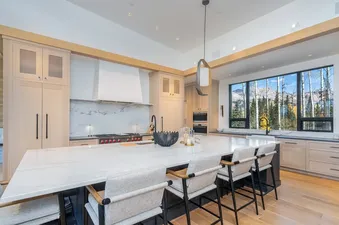 A modern mountain kitchen with light wood cabinetry, a large marble island, and a wide window showcasing a mountain view with pine and aspen trees.