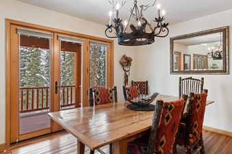 A rustic dining room with a wooden table and patterned chairs, illuminated by an antler-themed chandelier, with a view of a snow-covered forest through large glass doors.