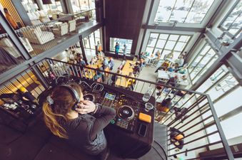 An elevated view of a DJ playing music for a lively crowd in the open-concept lounge at Gravity Haus JH Two Queens.
