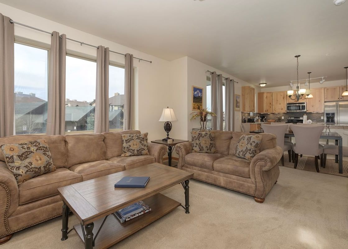 The living area of Trailhead Lodges 422 features two brown sofas with patterned throw pillows, a wooden coffee table, and large windows with cream-colored curtains.