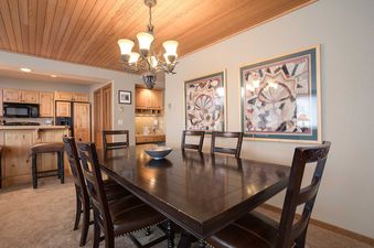 The dining room of Chateau Chamonix CX124 showcases a dark wood dining table with six chairs, an elegant chandelier, and framed artwork, seamlessly connected to a modern kitchen with light wood cabinetry.