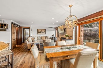 A spacious great room in Bear Claw 2 Valley View 513, featuring a dining area with a rustic wooden table and a living room with a sectional sofa and a flat-screen TV above a fireplace, all bathed in natural light.