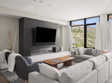 Modern living area of One Snowmass East 603 with comfortable seating, a large wall-mounted TV, and scenic mountain views through floor-to-ceiling windows.