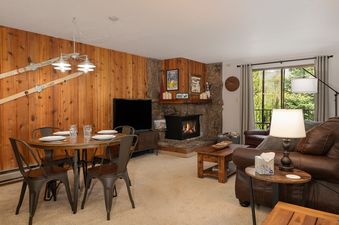 A cozy living room at Beaver Village 823 features a dining area with a round wooden table and metal chairs, a stone fireplace, and a comfortable brown leather sofa, with a large window offering a view of lush green trees.