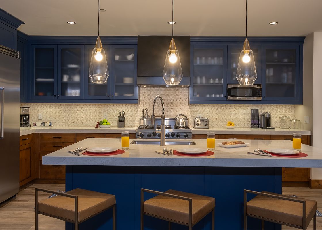 A modern kitchen with navy blue cabinetry, stainless steel appliances, a large island with breakfast settings, and elegant pendant lighting.