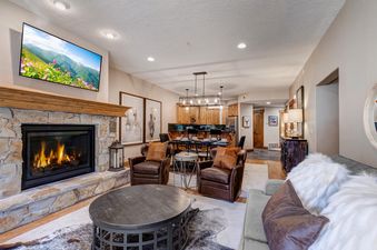A cozy, open-concept living room features a stone fireplace with a TV above, leather armchairs, a gray sofa, and a dark coffee table, leading into a dining area and kitchen.