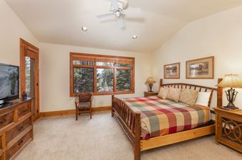 A cozy bedroom with a wooden bed frame, a colorful plaid comforter, and large windows overlooking a scenic forest.