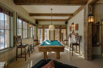 The games room at Elk Lake Lodge features a rustic pool table with a green felt surface, surrounded by wooden bar stools and large windows looking out onto a snowy landscape.
