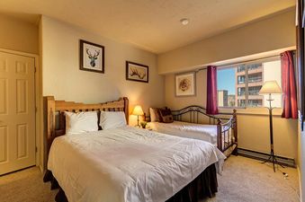A bedroom with two beds, one with a white bedspread and the other with a brown bedspread. There is a window with a view of the mountains in the background.