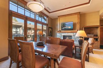 A well-lit dining room with a wooden table and chairs set against a large window offering an exterior view of a condo complex.