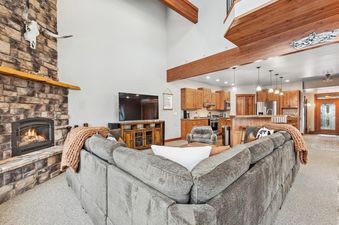A cozy living room in the Chair View Chalet featuring a large L-shaped sofa, a stone fireplace with a fire, a flat-screen TV, and an open-concept kitchen with wooden cabinetry and a breakfast bar.