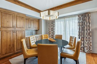 A formal dining area in the St Regis Executive Suite features a dark round table surrounded by six light brown leather chairs, with light wood cabinetry on the left wall and patterned curtains on the right.