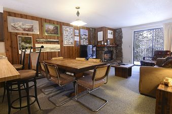 The dining and living area of Beaver Village 1821 features a wooden dining table with six chairs, a cozy fireplace, and a sliding glass door leading to a balcony with a view of snow-covered trees.