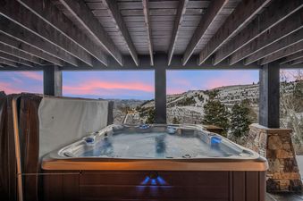 A steaming outdoor hot tub under a covered wooden deck at Mountains Edge overlooking a snowy mountain landscape at sunset.