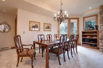 The Chateau Chamonix CX324 dining area features a large wooden dining table with six upholstered chairs, illuminated by a chandelier and natural light from a window and glass door revealing an outdoor view.