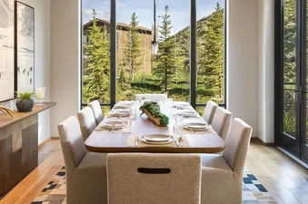A contemporary dining room with a wooden table set for eight, surrounded by upholstered chairs, and large windows offering views of green mountains and pine trees.