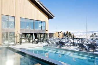 An inviting outdoor heated pool and hot tub area at Gravity Haus JH Two Queens during winter, with snow-covered mountains in the distance.