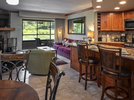 The living area of Building 4 One Bedroom Beaver Run Resort, featuring a stone fireplace, purple sofa, and a kitchen with wooden cabinetry and granite countertops, with a view of green foliage outside.