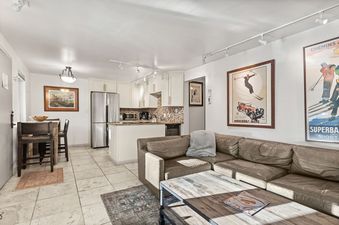 A bright open-concept living area with a tan leather sectional, rustic wooden coffee table, and an adjacent kitchen with cream cabinetry and stainless steel appliances.