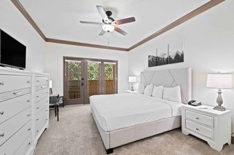 A bright and airy bedroom at The Charter E0205 with a large bed, white furniture, and a view of trees through multiple glass-paned doors.