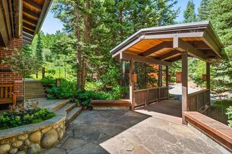A stone patio and a covered wooden walkway lead through a lush, forested exterior with tall pine trees surrounding the Lupine Estate.