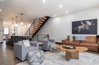 An inviting open-concept living room with a brown leather sofa, two gray armchairs, a live-edge coffee table on an area rug, and a modern kitchen with a dark island and pendant lights.