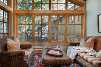 A living room with two wicker sofas, a coffee table with a book on it, and a large window with a view of trees and a forest.