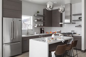 A modern kitchen with dark wood cabinets, white countertops, stainless steel appliances, and two spherical pendant lights, with a large window overlooking a snowy landscape.