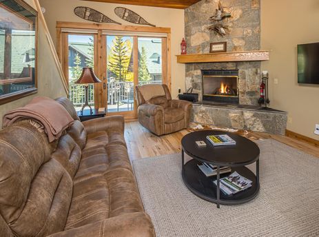 A cozy lounge area with a brown leather sofa, an armchair, a stone fireplace, and large glass doors leading to a balcony.