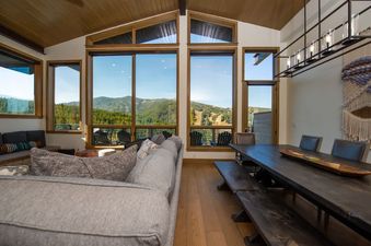 The living area and dining area of Apex Manor feature a large gray sectional sofa and a long wooden dining table with benches, all set against a backdrop of expansive windows showcasing a stunning mountain and forest view.