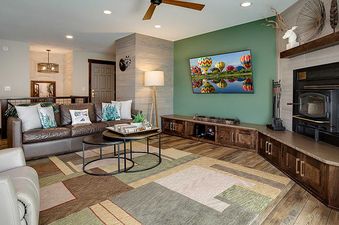 A modern and rustic living room and kitchen at The Ranch at Steamboat RA217 features a brown leather sofa, a flat-screen TV, dark wood cabinetry, and unique lighting, all set under a patterned ceiling.